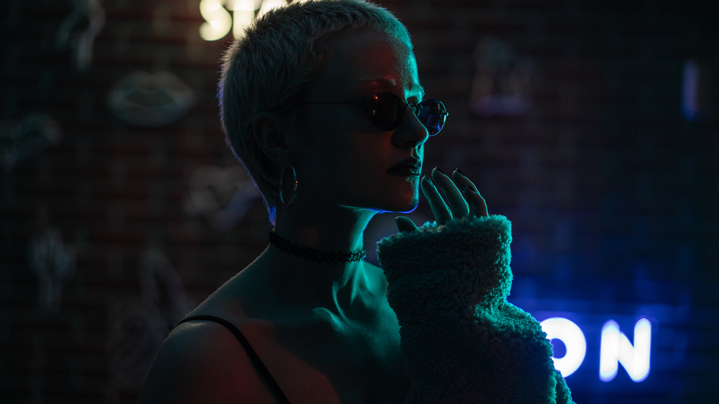 A woman with short cropped hair standing next to a brick wall with neon signs illuminated behind her. Despite the recession she is fashionably dressed with sunglasses and a fluffy jacket.