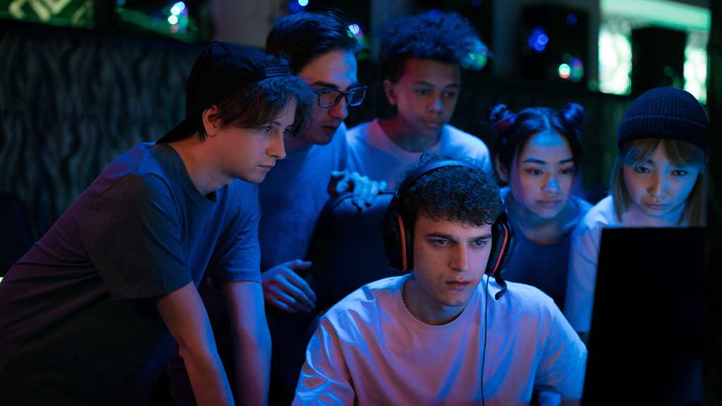 A number of young people surrounding one person playing games on Twitch, looking at the computer screen with interested but concerned expressions.