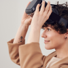 A woman with short hair and a baggy brown shirt holding up a VR headset on her head, similar to the Apple Vision Pro.