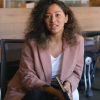 A young Black woman with curly hair sits in a chair and is talking to a person who stands off-screen despite imposter syndrome.