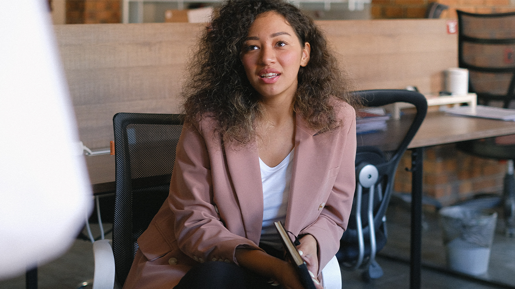 A young Black woman with curly hair sits in a chair and is talking to a person who stands off-screen despite imposter syndrome.