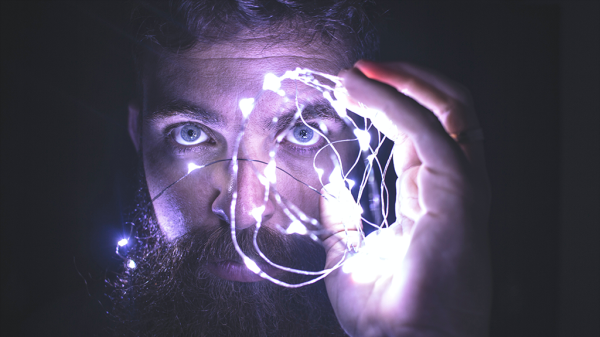 A bearded man holding up fairy lights up to his face to illuminate his face in a blue-purple light representing neurotechnology.