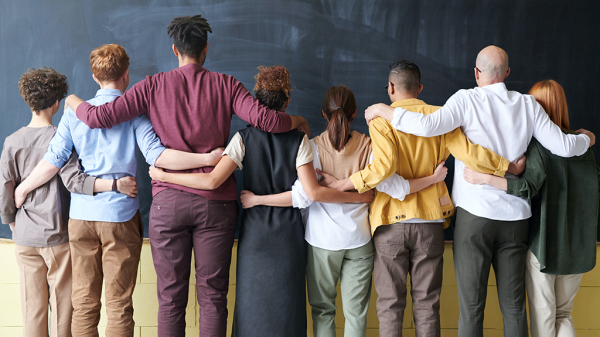 A varied group of people facing away from the camera and holding on to each other by shoulder and waist. The group showcases diversity. with a variety of skin tones, heights, and genders represented.