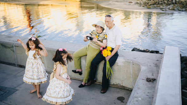An elderly couple film their grandchildren on their phone while they sit beside a body of water, showing a gap in spending habits for hobbies.