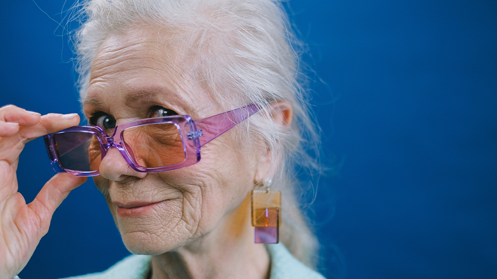 An older woman wearing purple glasses with tinted lenses and colorful earrings on a dark blue background with a mischievous smile following personality tests.