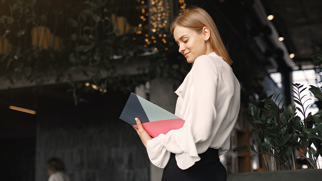 A blonde woman stands in a modern style cafe reviewing her entrepreneur planner and smiling toward her work.