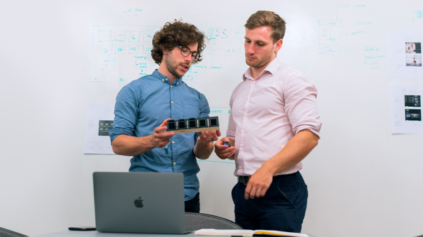 Two men stand next to a laptop, with one holding up a new product that the two discuss eagerly.