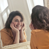 A woman with curly brown hair and a yellow shirt resting her hands on her chin as she smiles into a mirror, with our view looking over her shoulder at her personality.