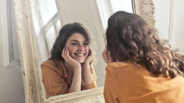 A woman with curly brown hair and a yellow shirt resting her hands on her chin as she smiles into a mirror, with our view looking over her shoulder at her personality.