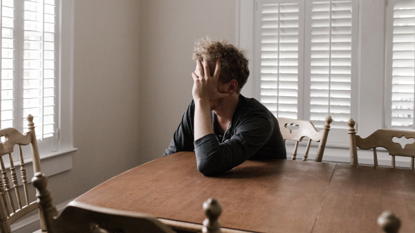 A person sitting at a kitchen table with their face obscured by their hands in the stress of rejection.