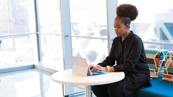 A Black woman sits at a comfortable seat with a laptop open next to a large office window working in her ADA accommodated space.