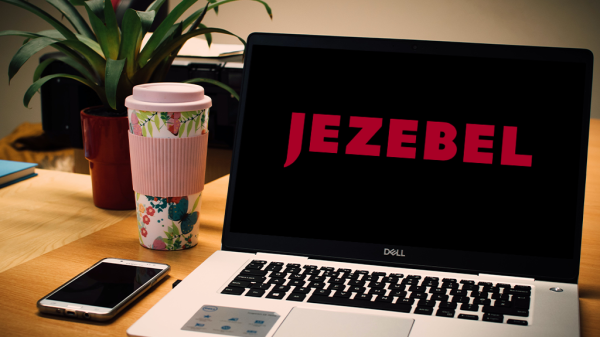 A laptop computer on a desk with a coffee and smart phone next to it. The logo for Jezebel, the news site, is pulled up on the screen.