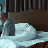 An older man sitting on the edge of a hotel bed with a heavy sad expression as he ponders an EEOC lawsuit.