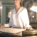 A woman standing at a hotel desk, with the focus on the foreground with the sign 'reception' illuminated by a frilled white desk lamp. The desk shows no ADA accommodation for the receptionist.