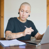 A woman with a bald head from cancer treatment writing and consulting a laptop for work.