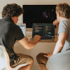 Two men, one with curly hair, and one with blonde hair, sit together with a laptop between them open to a coding page for the development of AI solutions.