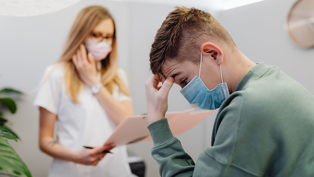 A man in a doctor's office, fingers on his forehead as he deals with the stress of health insurance, being filled out by a woman in the blurred background behind him.