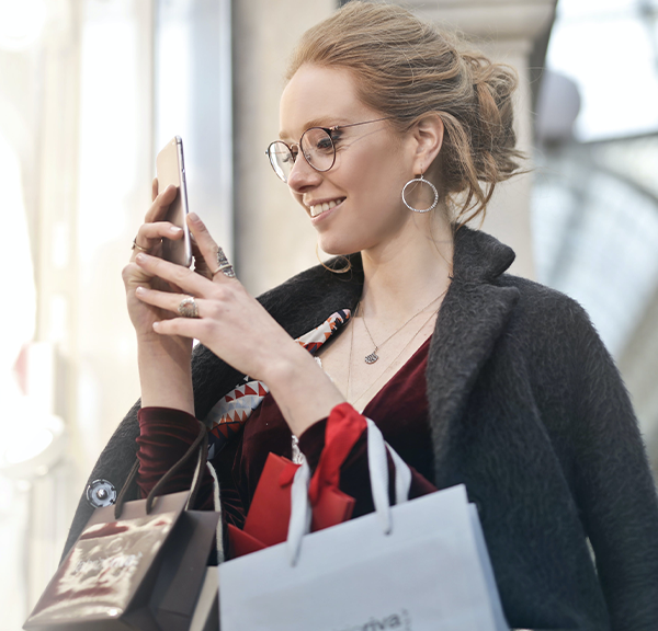 A woman checking her phone after a session of doom spending, with several shopping bags hanging off of her arms while she smiles.