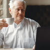 An older male employee contemplating retirement as he reads on his computer with a cup of coffee in one hand.