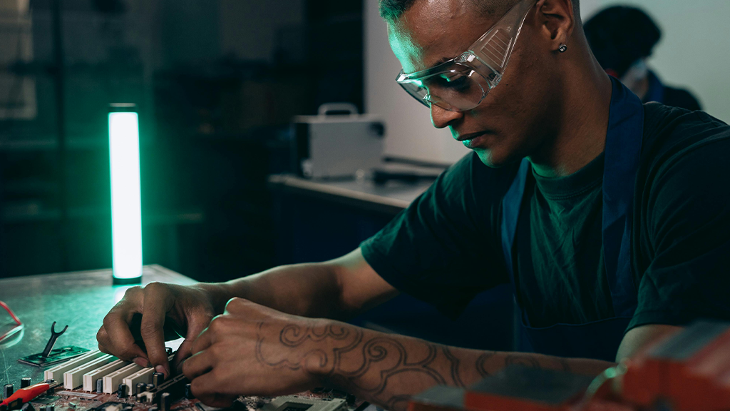 A Black man working on a computer microchip board, wearing safety goggles.