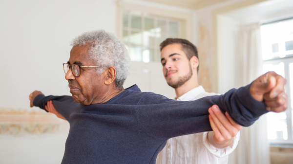 A younger man in healthcare work deserving of fair wages supporting an elderly Black man with his arms stretched out to either side.