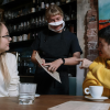 Two clients seated at a table speaking with a sign language interpreter to explain a document that she points at. She also wears a face mask with a clear window so lips can be read.