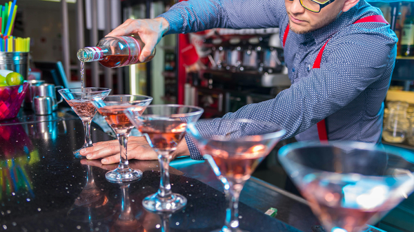 A person pouring red alcohol into a line of martini glasses on a counter, passing by a case for sexual harassment.
