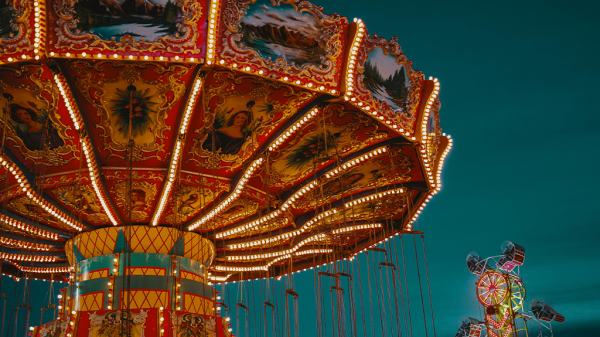 A night time view of a carnival ride illuminated in the twilight, manned by visa workers on the ground.