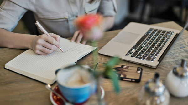 A person writes in a journal on a desk next to a cup of coffee, flowers, and an open laptop to compare to AI content.
