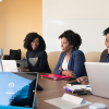 A group of Black female employees with different natural hair styles sitting at a business table with laptops, talking animatedly.
