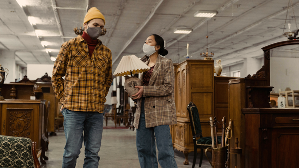 Two people in the middle of a furniture shop with unlawful rules around them. Both people are wearing face masks, and one holds a lamp as they talk to the other.
