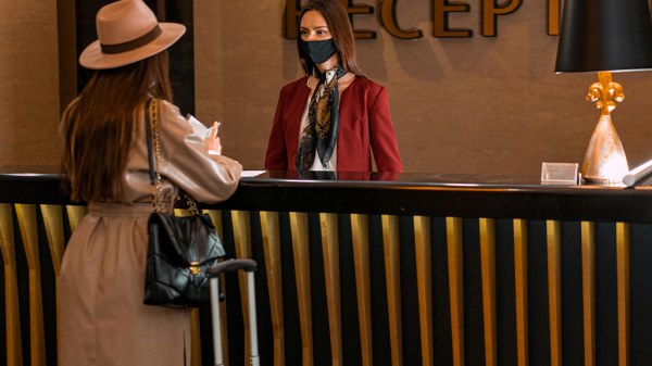 Two women wearing masks standing by a reception desk in a modern hotel with warm yellow lighting, with a sign against disability discrimination.