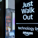 A close up view of a grab and go shopping area with a sign reading Just Walk Out, technology by Amazon on a black sign.