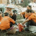 A group of migrant visa workers seated on a lawn during a break wearing orange shirts and talking as they eat.