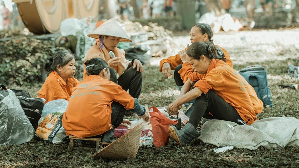 A group of migrant visa workers seated on a lawn during a break wearing orange shirts and talking as they eat.