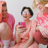 Three people in pink outfits, including one with pink hair, looking at a pink smartphone open to Airchat.