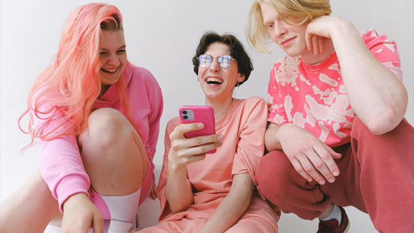 Three people in pink outfits, including one with pink hair, looking at a pink smartphone open to Airchat.