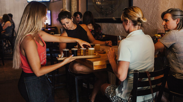 A woman serving beers in a restaurant to other patrons at a table for tips.