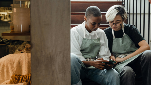 Two restaurant workers sitting together in a stairwell, one Black woman and one Asian woman comparing information on their phones about sexual harassment and how to combat it.