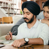 A man in a turban taps on a calculator with a laptop in front of him. A woman leans over his shoulder with some concern as they work on their tax deductions.