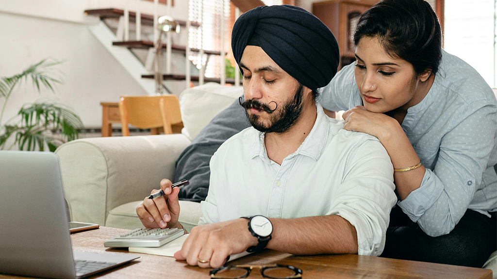 A man in a turban taps on a calculator with a laptop in front of him. A woman leans over his shoulder with some concern as they work on their tax deductions.