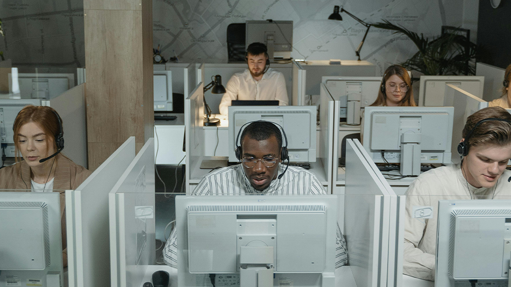 An office of call center customer service workers in small cubicles with headphones and talking to customers on the phone.