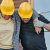 Two workers in construction hats, one worker holding onto the other as they tend to an injury off screen.