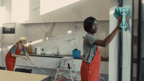 Two female janitors are cleaning an office kitchen. A Black woman in the foreground wipes down the window and a White woman in the back mops the floor. Child labor laws protect children from these hazardous chemicals.