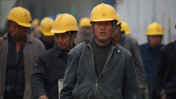 A group of miners with hard hats and dour expressions walk toward the camera, away from a company experiencing bankruptcy.