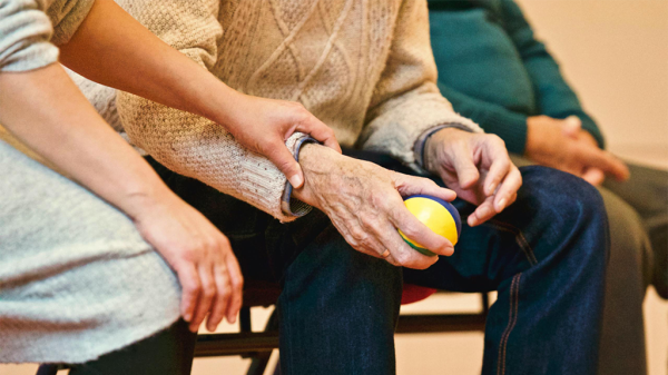 An older person with a supportive stress ball in one hand and having a second person hold their hand in nursing care, supported by OSHA rules.