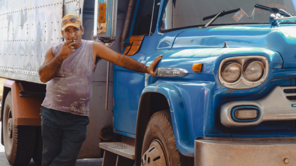A man smokes a cigarette and leans up against a blue semi truck with a serious expression and hanging decoration in their truck protected by religious accommodations.